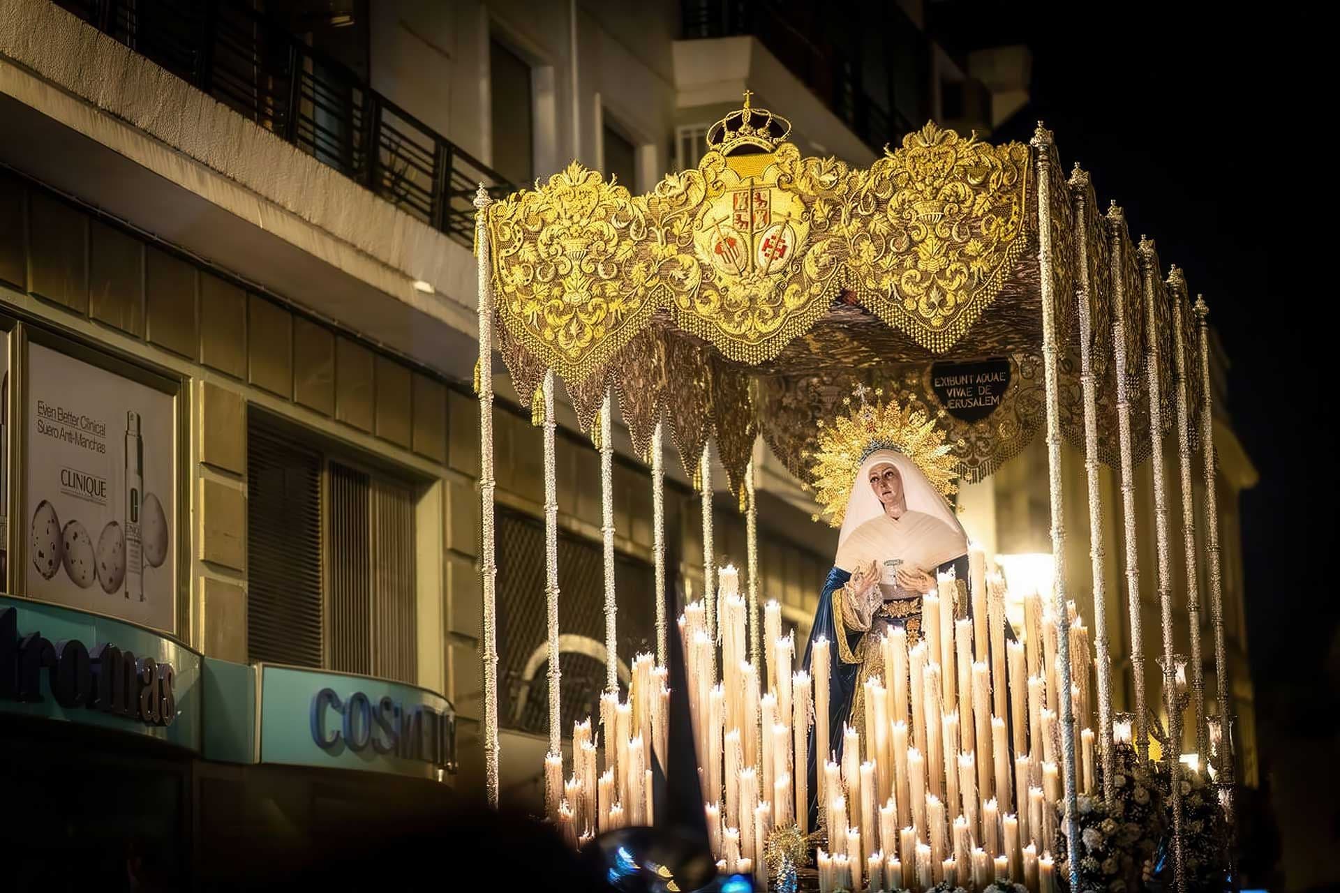A illuminated procession through Seville at night, with penitents in hooded gowns carrying an ornate image of the Virgin Mary surrounded by flickering lanterns.